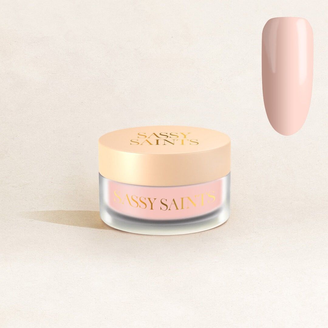 A small frosted jar with a beige lid labeled Sassy Saints - Brings The Salon Home Peachy Pearl Dipping Powder sits on a neutral background, with a glossy pale nude-pink swatch shown in the top right corner.