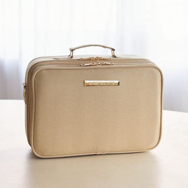 The Sassy Saints - Brings The Salon Home Large Storage Box is a beige, rectangular organizer with a top handle, gold zippers, and a front gold nameplate, placed on a light surface with sheer curtains in the background.