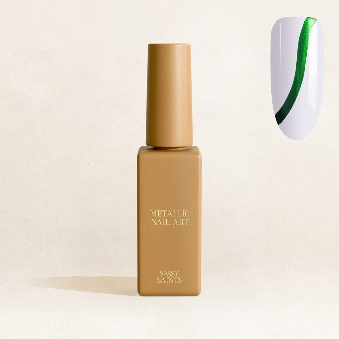 A gold bottle labeled Emerald Envy Metallic Nail Art Polish by Sassy Saints - Brings The Salon Home, next to a nail design with two curved metallic green lines on a white background.