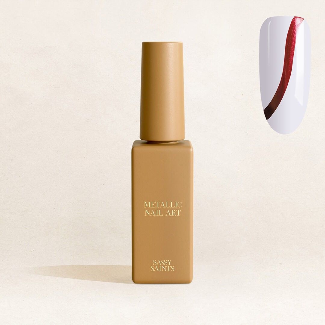 A golden bottle of Sassy Saints - Brings The Salon Home Ruby Rockstar Metallic Nail Art Polish stands upright on a light surface. To the right, a nail displays a diagonal red and brown metallic design on glossy white.