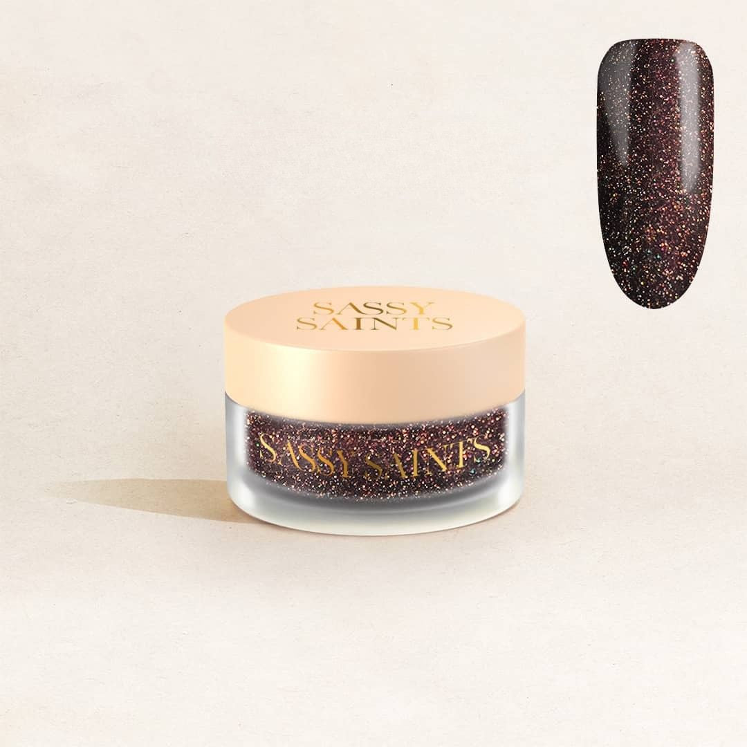 A small round jar labeled Sassy Saints - Brings The Salon Home with a beige lid, containing Cosmic Charmer Dipping Powder. A swatch above shows a shiny deep brown shade with fine golden and red sparkles.