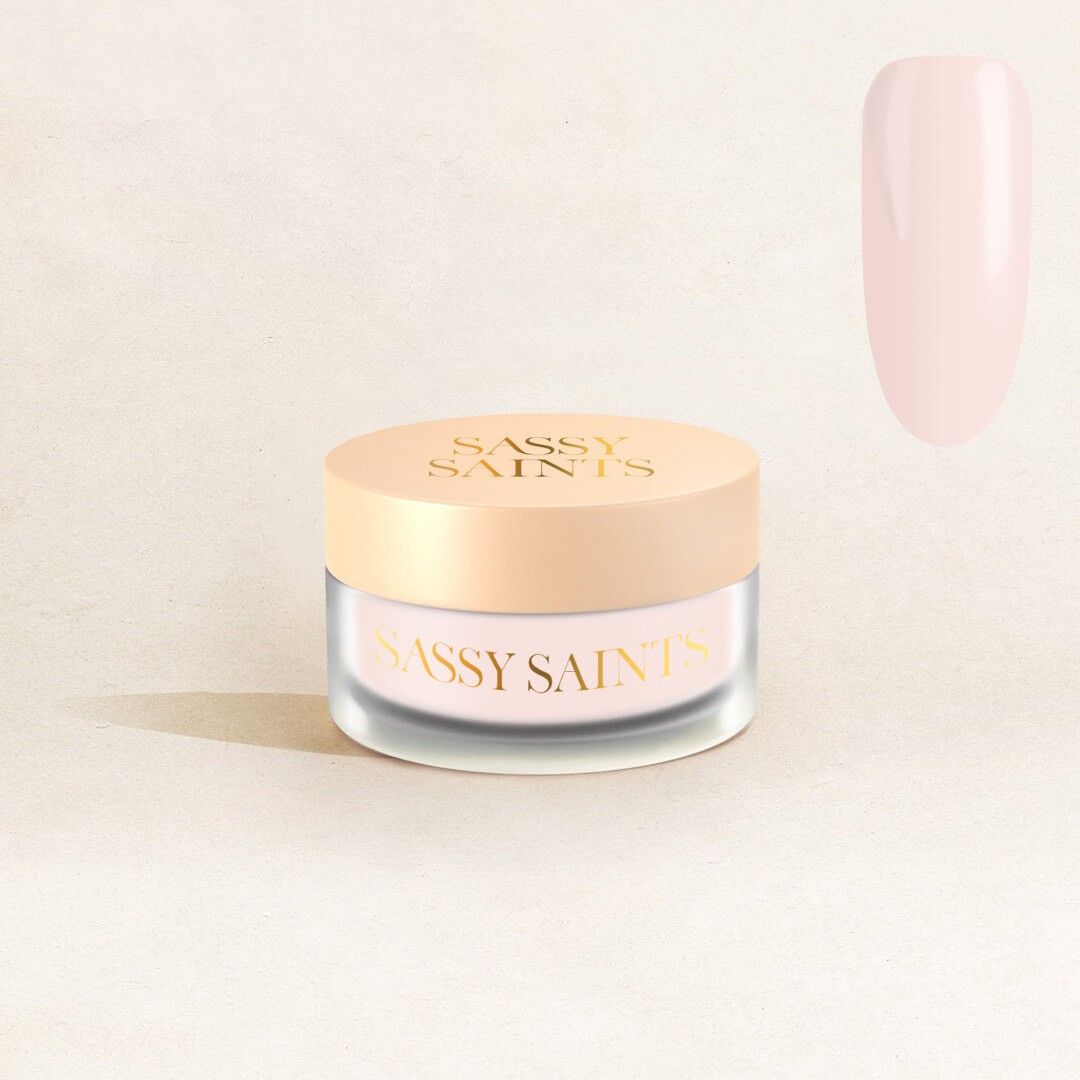 A small frosted glass jar with a beige lid labeled "Soapy Sweetness Dipping Powder" by Sassy Saints - Brings The Salon Home sits on a light beige surface, with a vertical swatch of pale pink nail powder to its right.