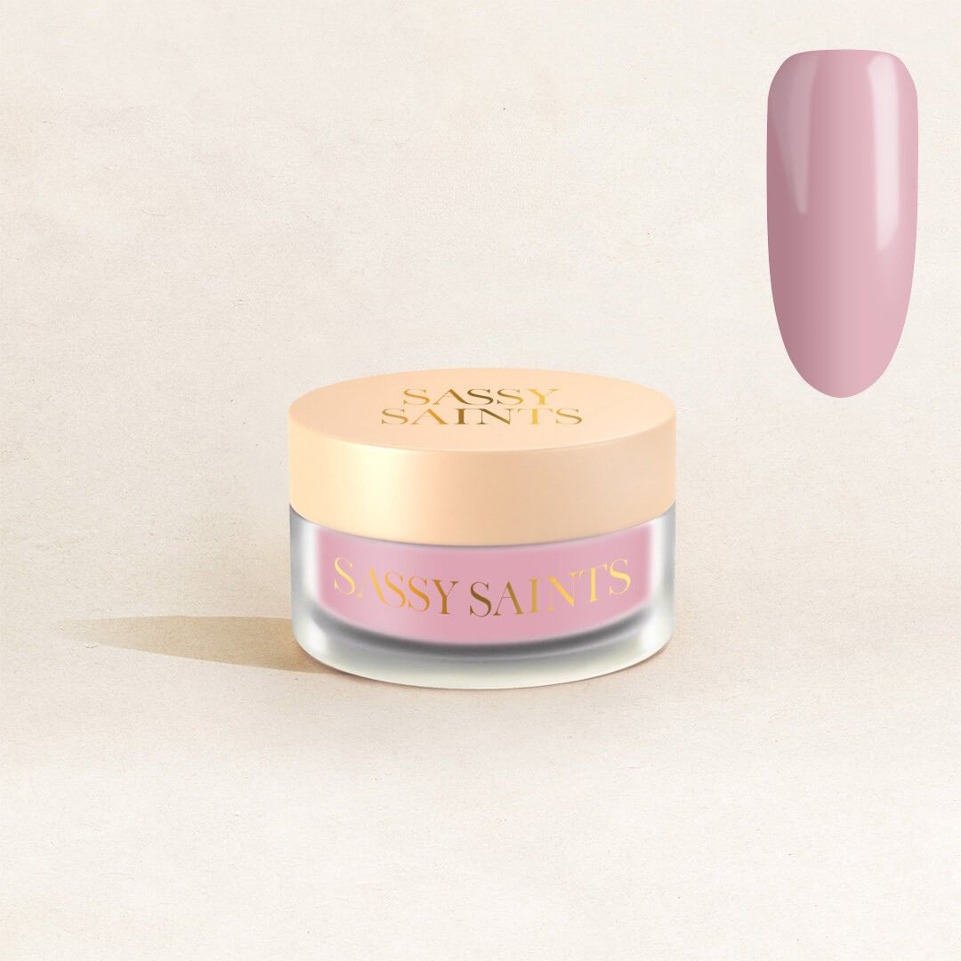 A small glass jar with a beige lid labeled "Sassy Saints - Brings The Salon Home" sits on a light background. Above it, a large swatch shows glossy mauve-pink, matching the Petal Pink Dipping Powder inside.