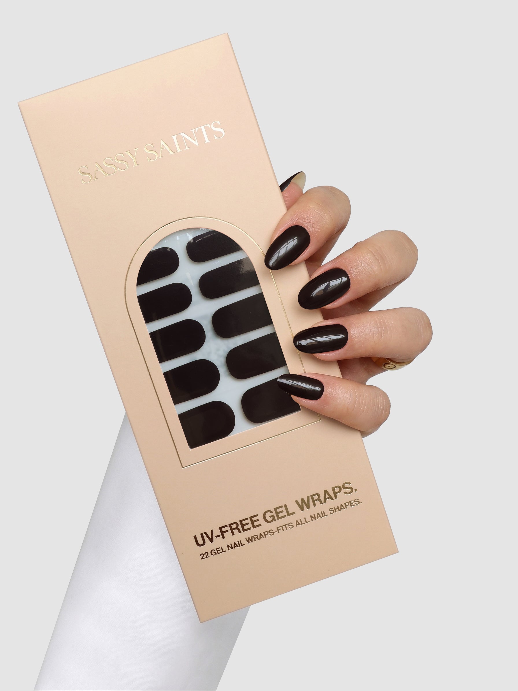 A hand with dark, glossy nails holds a beige box labeled “Sassy Saints – Brings The Salon Home,” containing Velvet Vice Gel Wrap UV-free nail wraps in a dark shade. The box features a clear window displaying the wraps inside.
