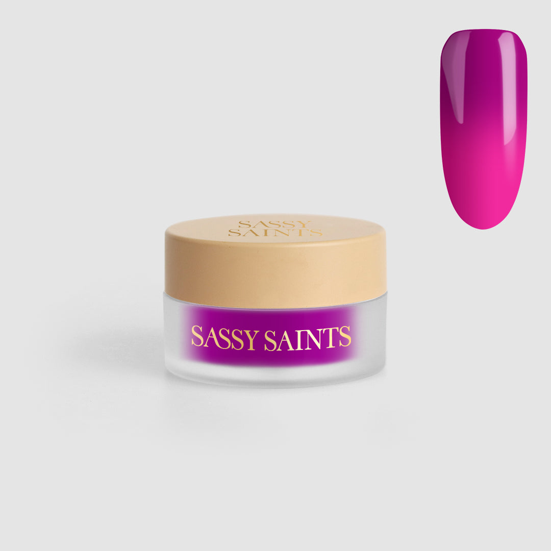 A small jar of Sassy Saints - Brings The Salon Home Vivid Voltage Dipping Powder with a beige lid and bright magenta powder sits beside a nail swatch painted in glossy magenta.