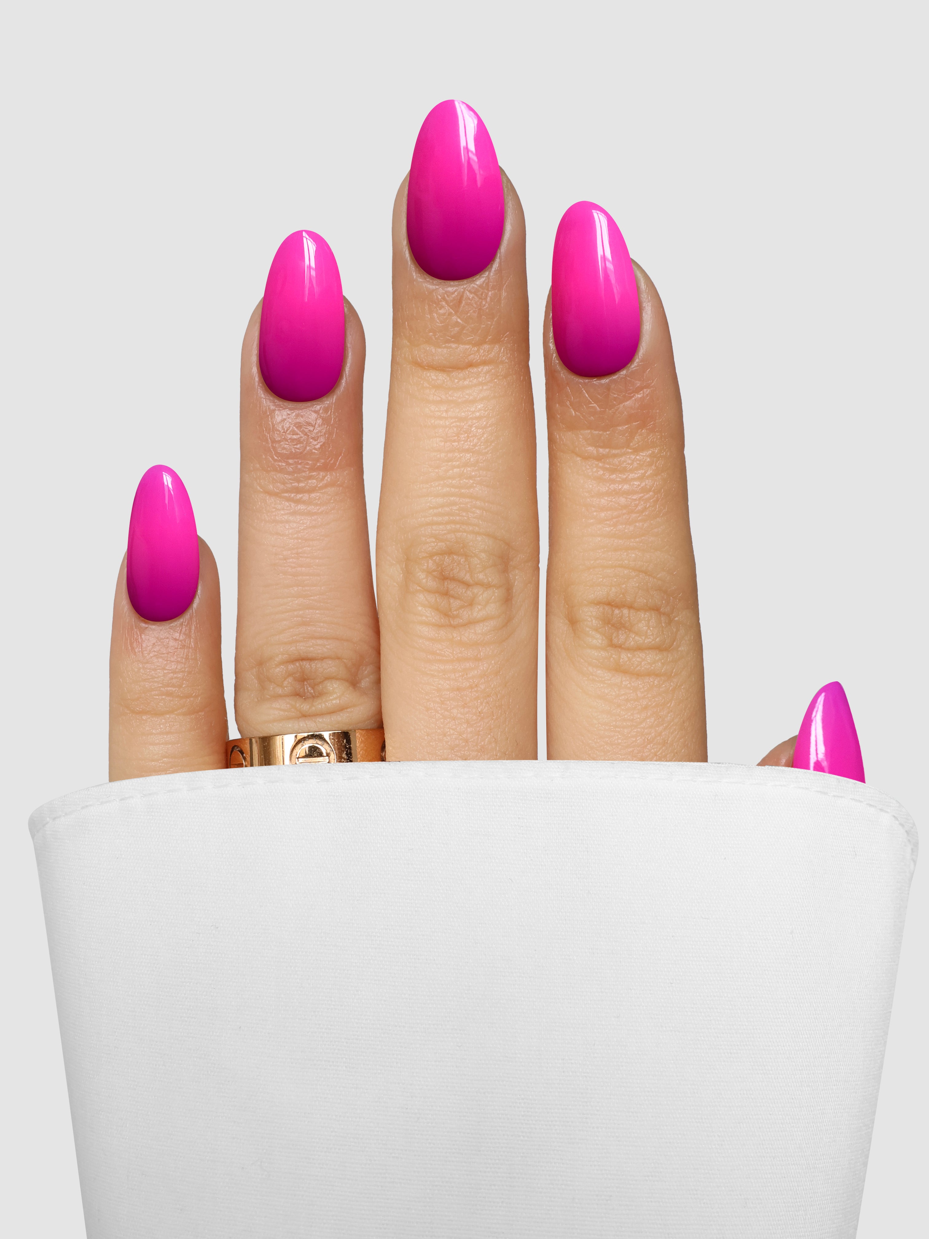 A hand with bright pink, almond-shaped nails created using Sassy Saints Vivid Voltage Dipping Powder peeks from a white sleeve, showcasing a gold ring on the middle finger against a plain light grey background.