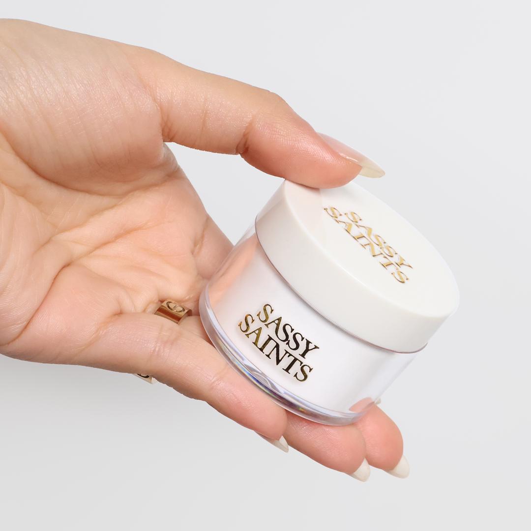 A hand with manicured nails holds a white Clear Base Vitamin Powder 30g jar from Sassy Saints – Brings The Salon Home, set against a plain, light background.