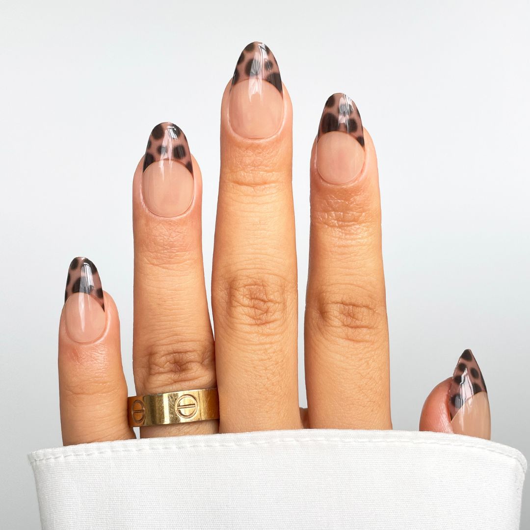 A hand displays Sassy Saints' Wild French Gel Wraps—almond-shaped nails with a nude base and leopard print tips—plus a gold ring on the middle finger and a white cuff partly covering the wrist.