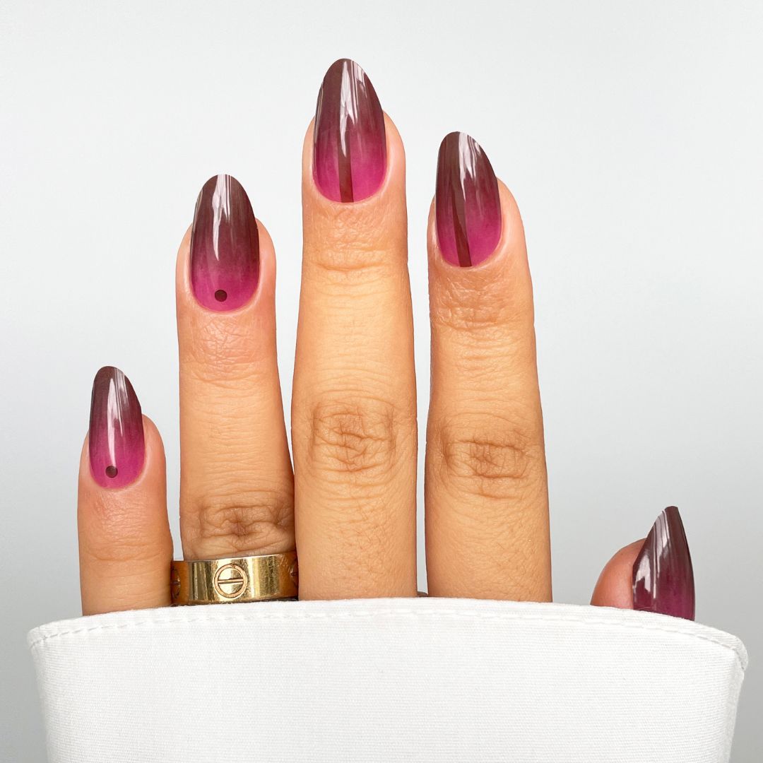 A hand with long almond-shaped nails in Cherry Smoke Gel Wrap by Sassy Saints—featuring a gradient purple-pink design with a black dot at each base—partially covered by a white sleeve, and wearing a gold ring on the middle finger.