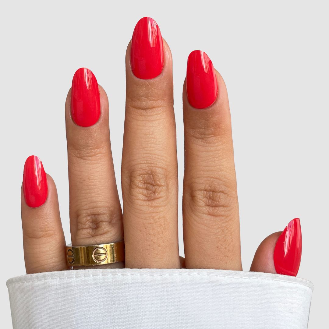 A hand with medium-length, oval nails painted bright red using Sassy Saints - Brings The Salon Home Hotline Edit Gel Wrap. The middle finger features a gold screw-design ring, partially covered by a white sleeve.