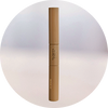 A brown, double-ended makeup stick stands upright against a soft, light background. The product features minimalistic gold text and a sleek cylindrical design.