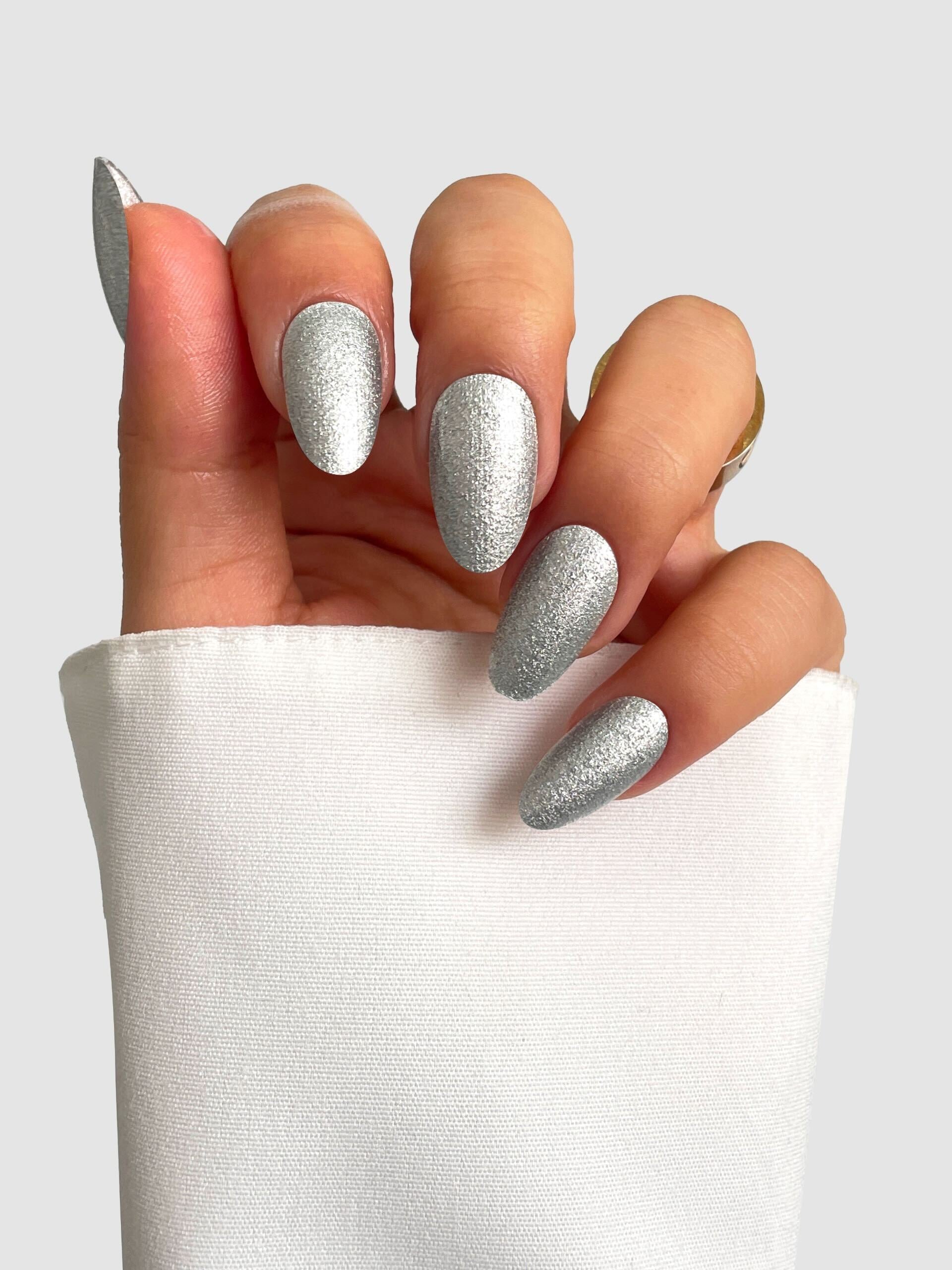 A hand with long, almond-shaped nails painted in Sassy Saints - Brings The Salon Home’s Liquid Platinum Gel Wrap, partially covered by a white sleeve, against a plain light gray background.