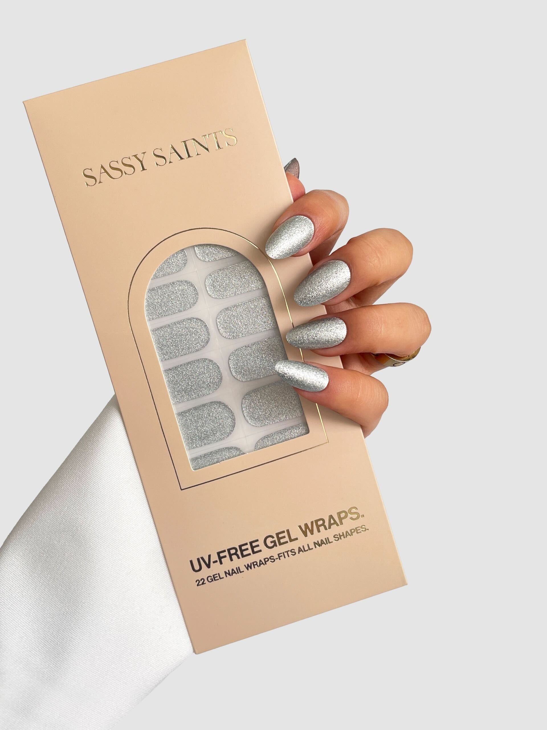 A hand with silver glittery gel nails holds a beige box labeled “Sassy Saints – Brings The Salon Home” Liquid Platinum Gel Wrap, with silver wraps visible through a window. The background is light gray.