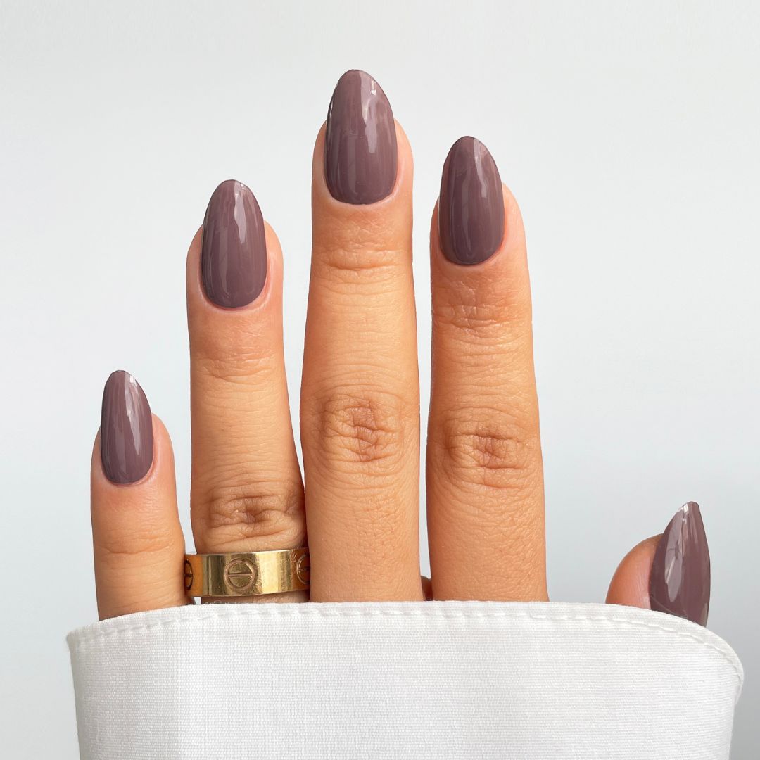 A hand with almond-shaped, glossy mauve Mocha Blend Gel Wrap nails by Sassy Saints - Brings The Salon Home, and a gold ring on the ring finger, is shown against a light background, partly covered by a white cuff.
