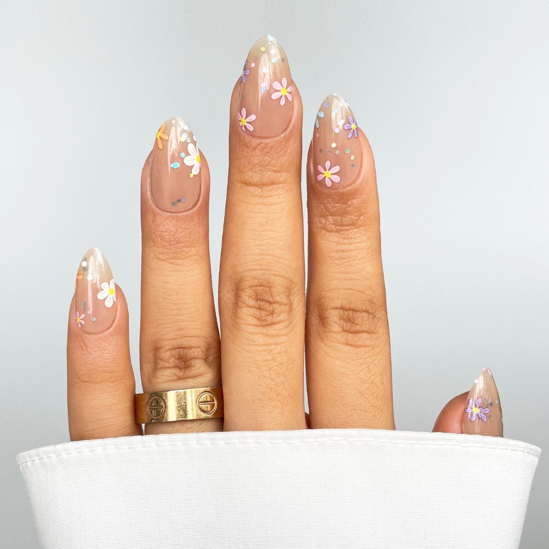 A hand showcases Sassy Saints - Brings The Salon Home's Petal Verse Gel Wrap: almond-shaped nails with a clear base, white tips, colorful floral accents, and dots. Two gold rings adorn the fingers; a white sleeve partially covers the hand.