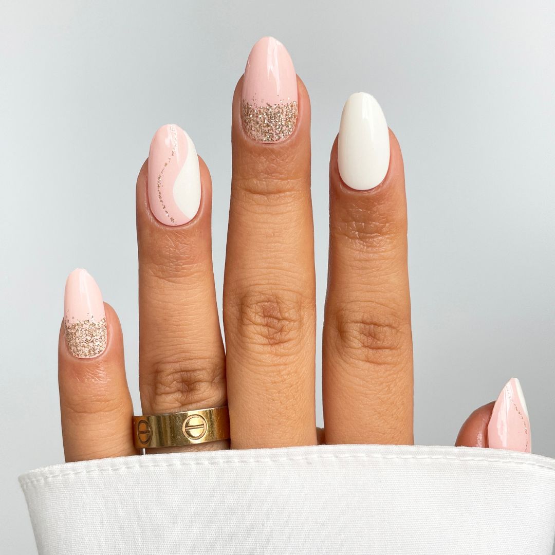 A hand with almond-shaped nails in Soft Launch Gel Wrap by Sassy Saints - Brings The Salon Home, showcasing a pink and white design with gold glitter accents and a gold ring, partially covered by a white sleeve on a light background.