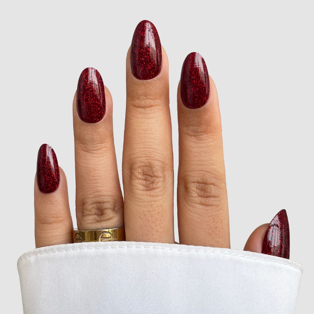 A hand with long, almond-shaped nails features Sparkling Ember Gel Wrap by Sassy Saints - Brings The Salon Home, a dark red glitter look. A gold ring adorns the ring finger, and a white fabric sleeve partially covers the hand.